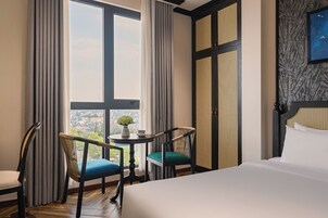 Une chambre d’hôtel avec un lit, deux chaises, une petite table avec un vase et de grandes fenêtres avec des rideaux.