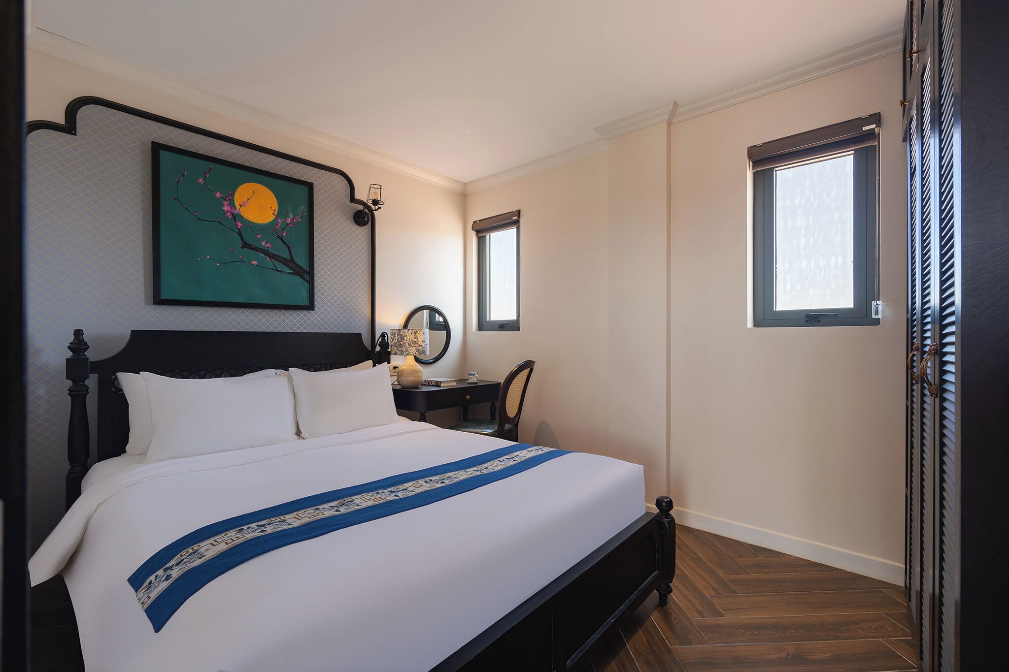 Una cama bien hecha con colcha azul y blanca, una mesita de noche con una lámpara, una pequeña mesa con un espejo y dos ventanas con persianas.