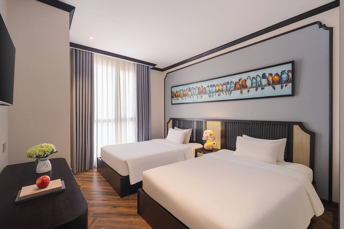 A hotel room with two beds, a flat-screen TV, a painting of birds, a vase with flowers, and a wooden floor.