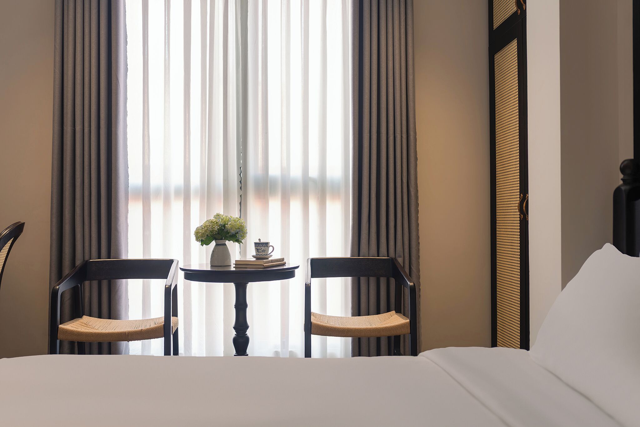 A hotel room with a bed, two chairs, a small table with a vase and books, and a window with curtains.