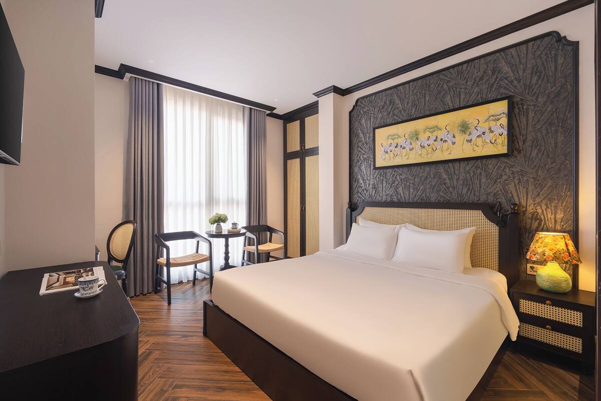 A hotel room with a large bed, a desk with a chair, a small table with chairs, and a painting on the wall.