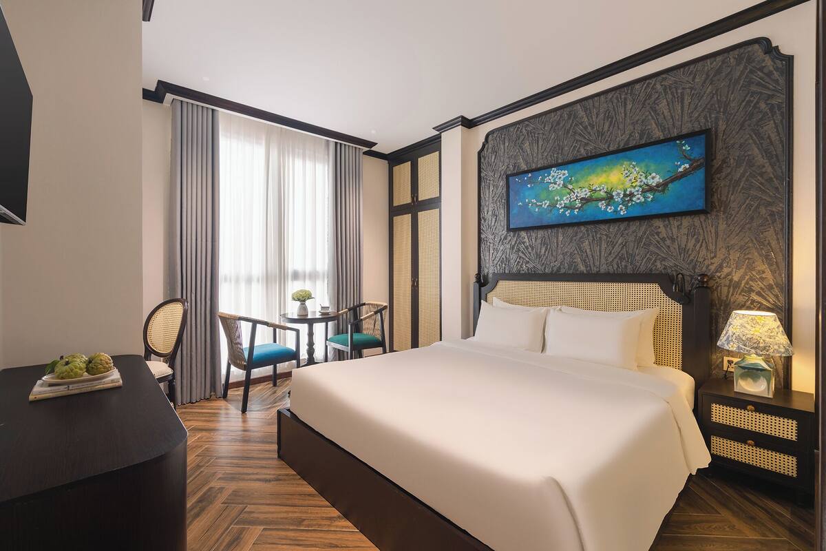 A hotel room with a large bed, a desk with a fruit tray, a chair, a small table, and a painting on the wall.