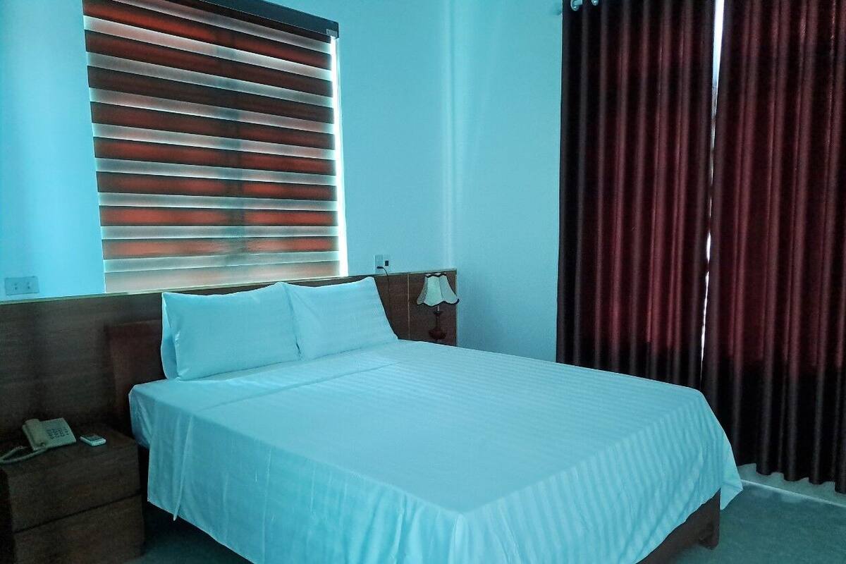 Een net opgemaakt bed in een hotelkamer met een houten hoofdbord, telefoon op het nachtkastje en een raam met blinds.