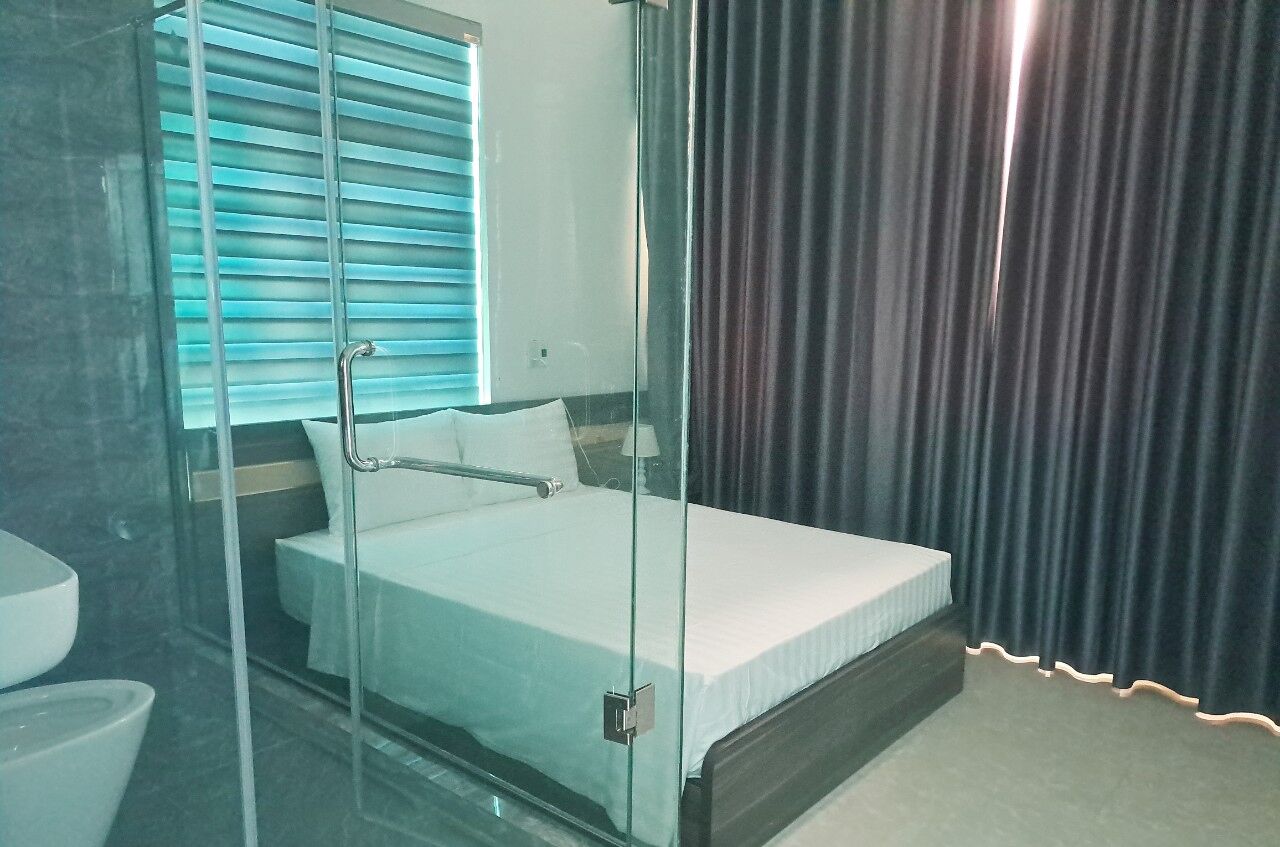 A hotel room with a glass-enclosed bed, a bathroom with a toilet and sink, and a window with blinds.