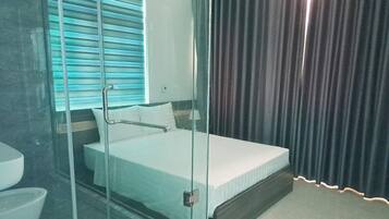 A hotel room with a glass-enclosed bed, a bathroom with a toilet and sink, and a window with blinds.