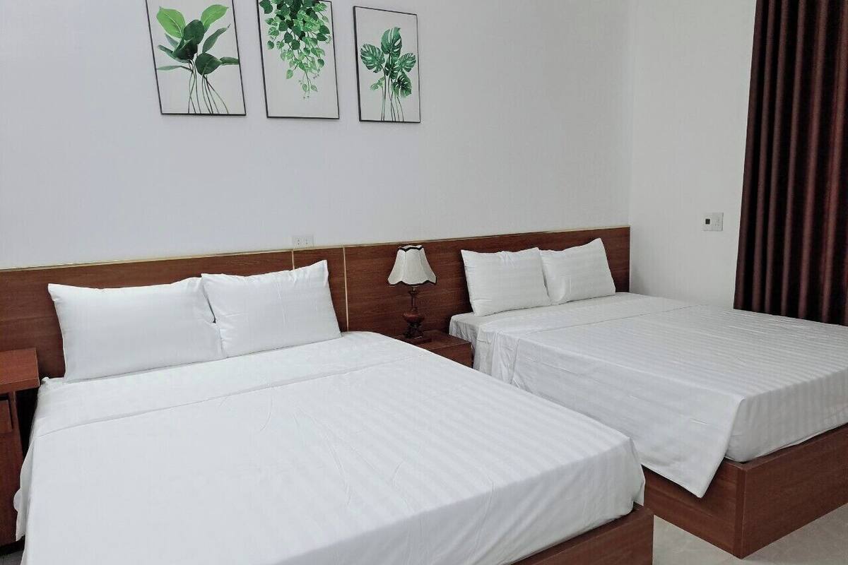 Een hotelkamer met twee enkele bedden, een houten hoofdbord, wit beddengoed, een bedlampje en drie lijstjes met plantenkunstwerken aan de muur.