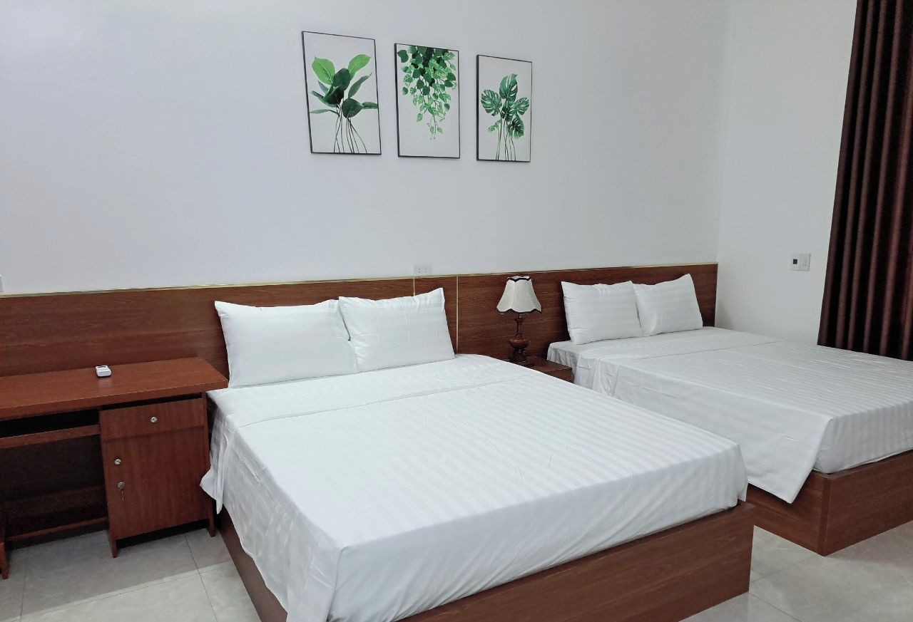 A hotel room with two beds, a wooden desk, and a nightstand. There are three framed botanical prints on the wall.