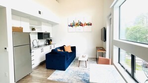 Un espacio habitable compacto con kitchenette, un sofá azul, una pequeña mesa y un ventanal con vistas al exterior.