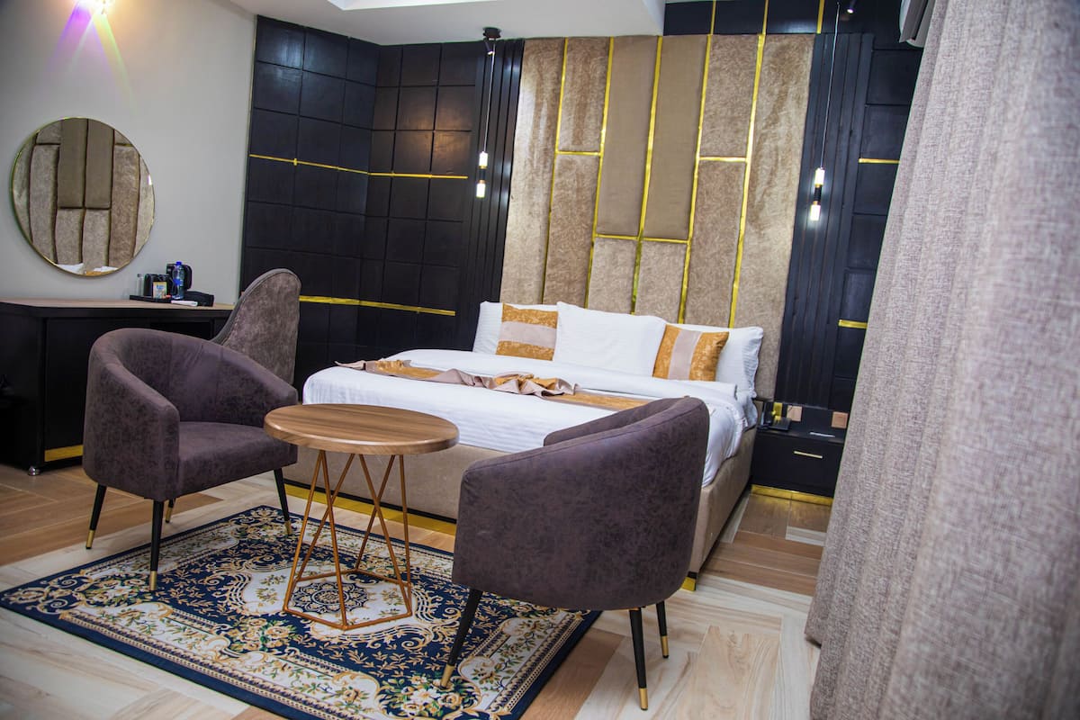 A modern bedroom with a large bed, two armchairs, a small round table, and a decorative rug.
