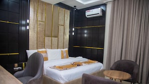 A modern hotel room with a large bed, two armchairs, a small wooden table, and a ceiling-mounted air conditioner.