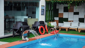 A swimming pool with a water feature, surrounded by lounge chairs and a sofa.