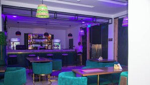 A bar area with teal chairs and tables, a bar counter with a variety of bottles, and a person standing behind the bar.