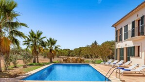 Una piscina con sillas de descanso y una mesa, rodeada de palmeras y un edificio con persianas.
