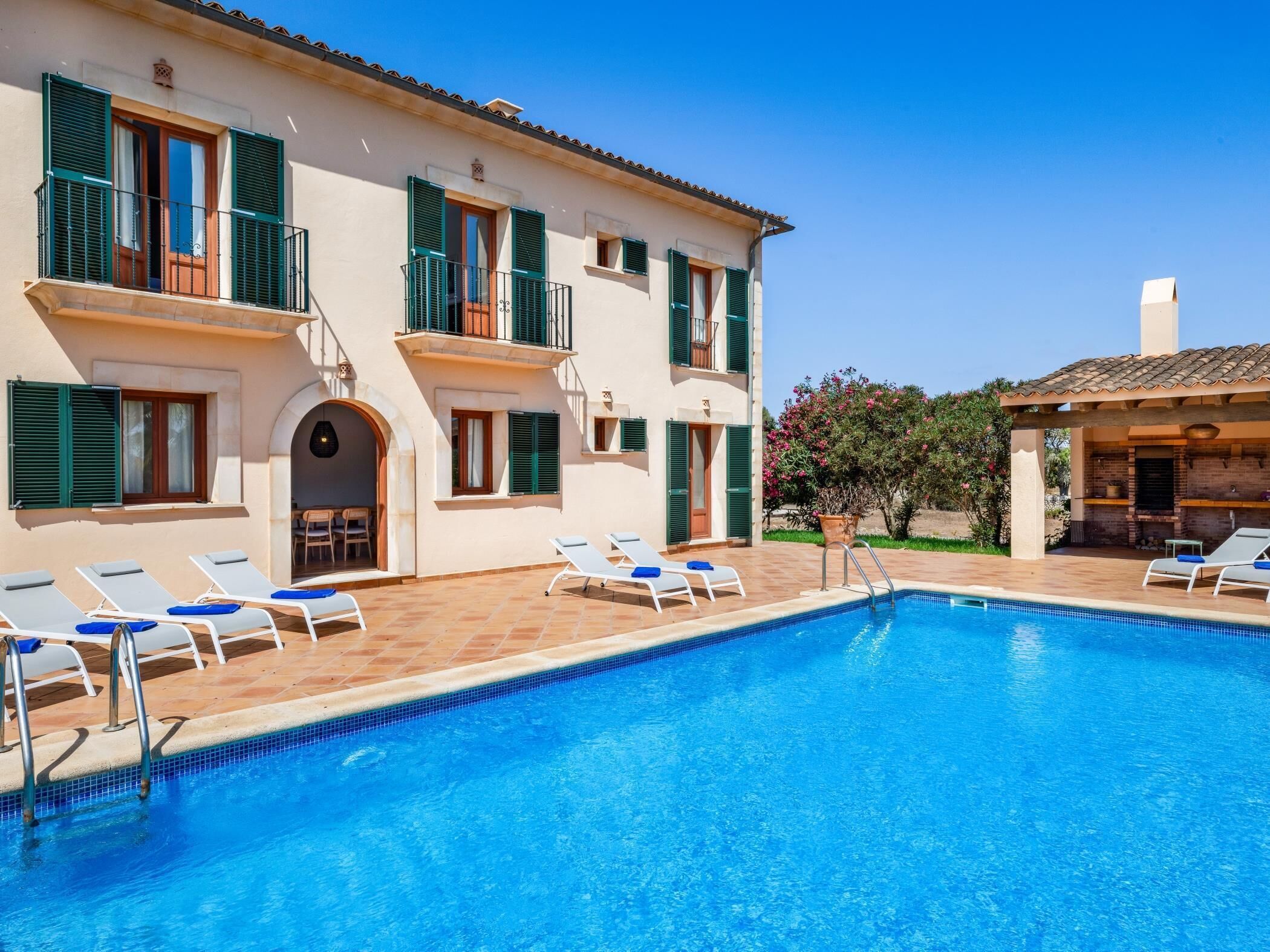 A poolside view of a building with a swimming pool, sun loungers, and a covered outdoor area.