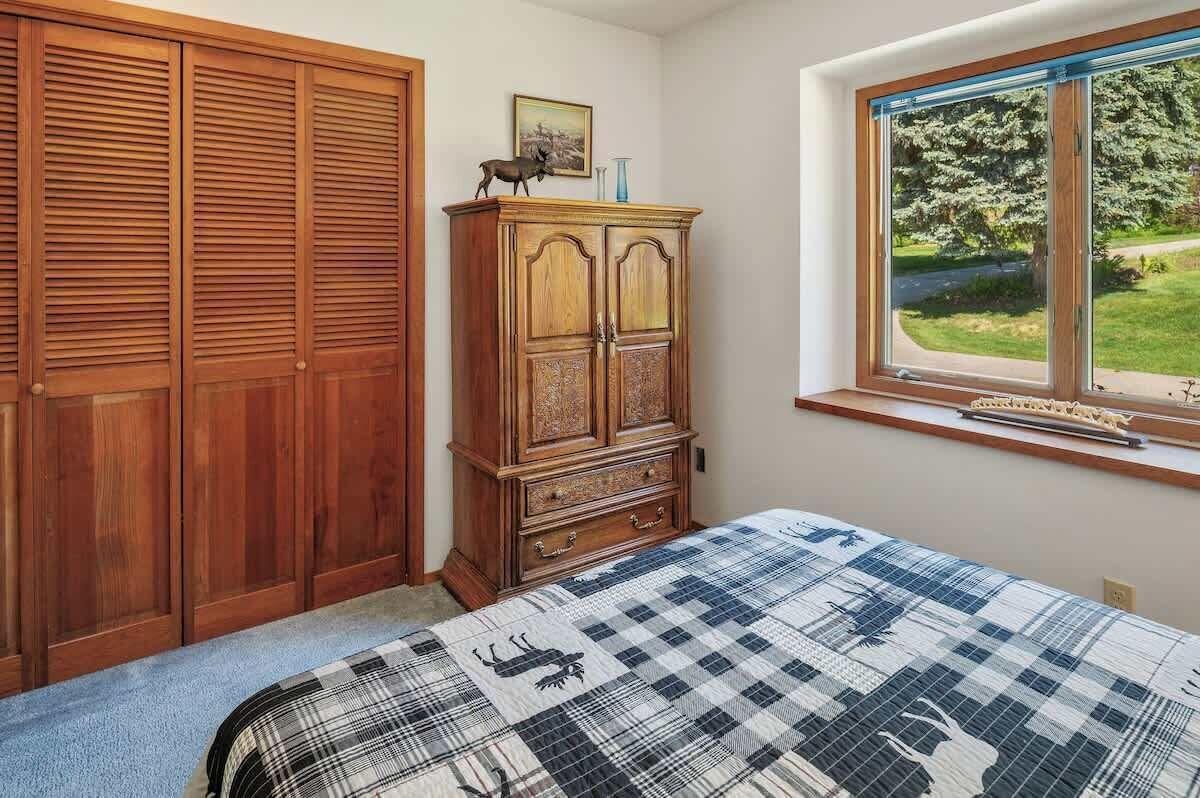 Een slaapkamer met een bed, een houten garderobe, een kast en een raam met uitzicht op groen.
