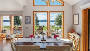 Un coin repas avec une table dressée pour quatre personnes, de grandes fenêtres offrant une vue sur un lac et des montagnes, et un lustre au-dessus.