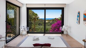 Une chambre avec une grande porte-fenêtre coulissante donnant sur un balcon, un lit agrémenté d’un coussin rouge et deux chaises blanches.