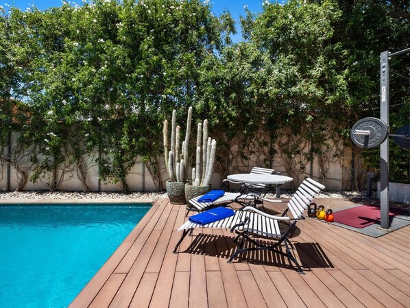 A poolside area with a wooden deck, lounge chairs, a table, and a weight bench.