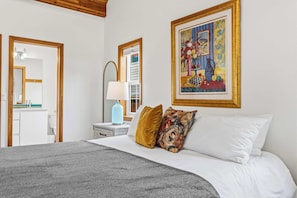 A bedroom with a bed, pillows, a nightstand, a lamp, a mirror, and a painting on the wall.