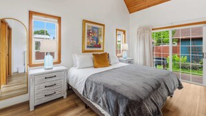 Um quarto com cama, mesas de cabeceira, cômoda, janela com cortinas e um quadro na parede.