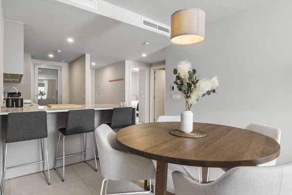 A modern kitchen with a dining area, featuring a round wooden table, grey chairs, and a vase with white flowers.