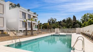 A swimming pool with a clear blue water, surrounded by a white fence, and a building with balconies and palm trees in the background. - Cosy Beach | Beach - pool - terrace (Marbella)
