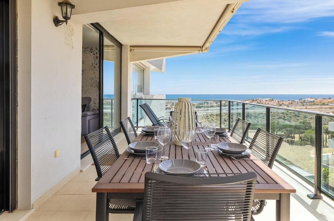 A dining table set on a balcony with a clear view of the sea and a distant town.