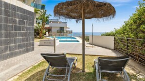 Ein Poolbereich mit zwei Liegestühlen, einem Reetdach-Sonnenschirm und Blick auf das Meer.