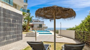 Un espace piscine comprenant deux chaises longues, un parasol en chaume et une vue sur la mer.