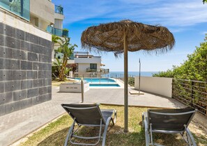 Un espace piscine comprenant deux chaises longues, un parasol en chaume et une vue sur la mer.