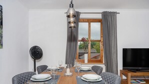 Een eetgedeelte met een houten tafel voor vier personen, een ventilator, een televisie en een raam met gordijnen.