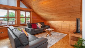 A loft-style room with a wooden ceiling, a sofa, a television, and a window with a view of greenery.