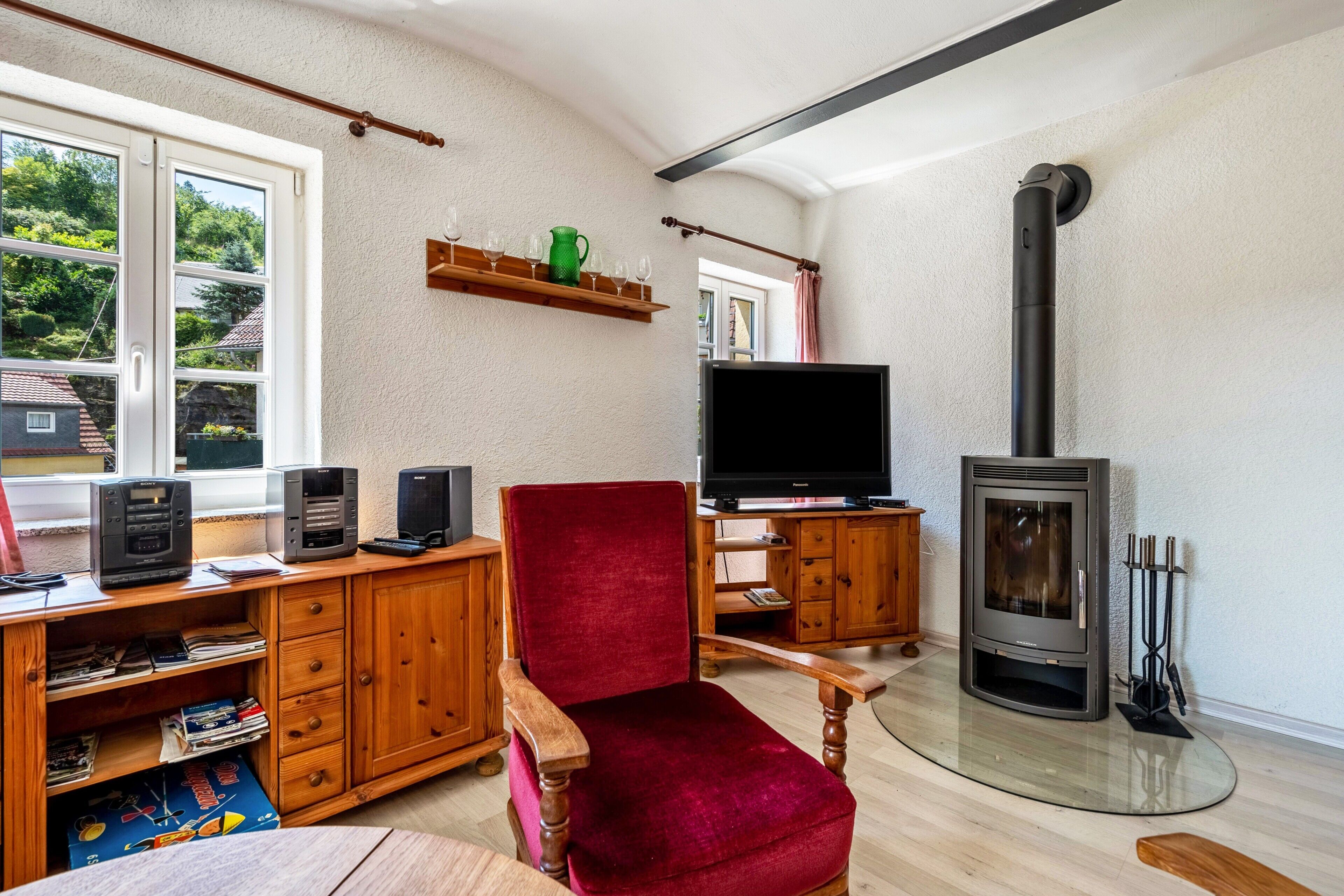 A living room with a TV, a red chair, a wooden desk, a fireplace, and a window with a view of trees.