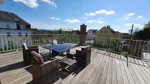 Eine Holzterrasse mit einem Tischtennis-Tisch, Korbmöbeln und einem Metallgeländer.