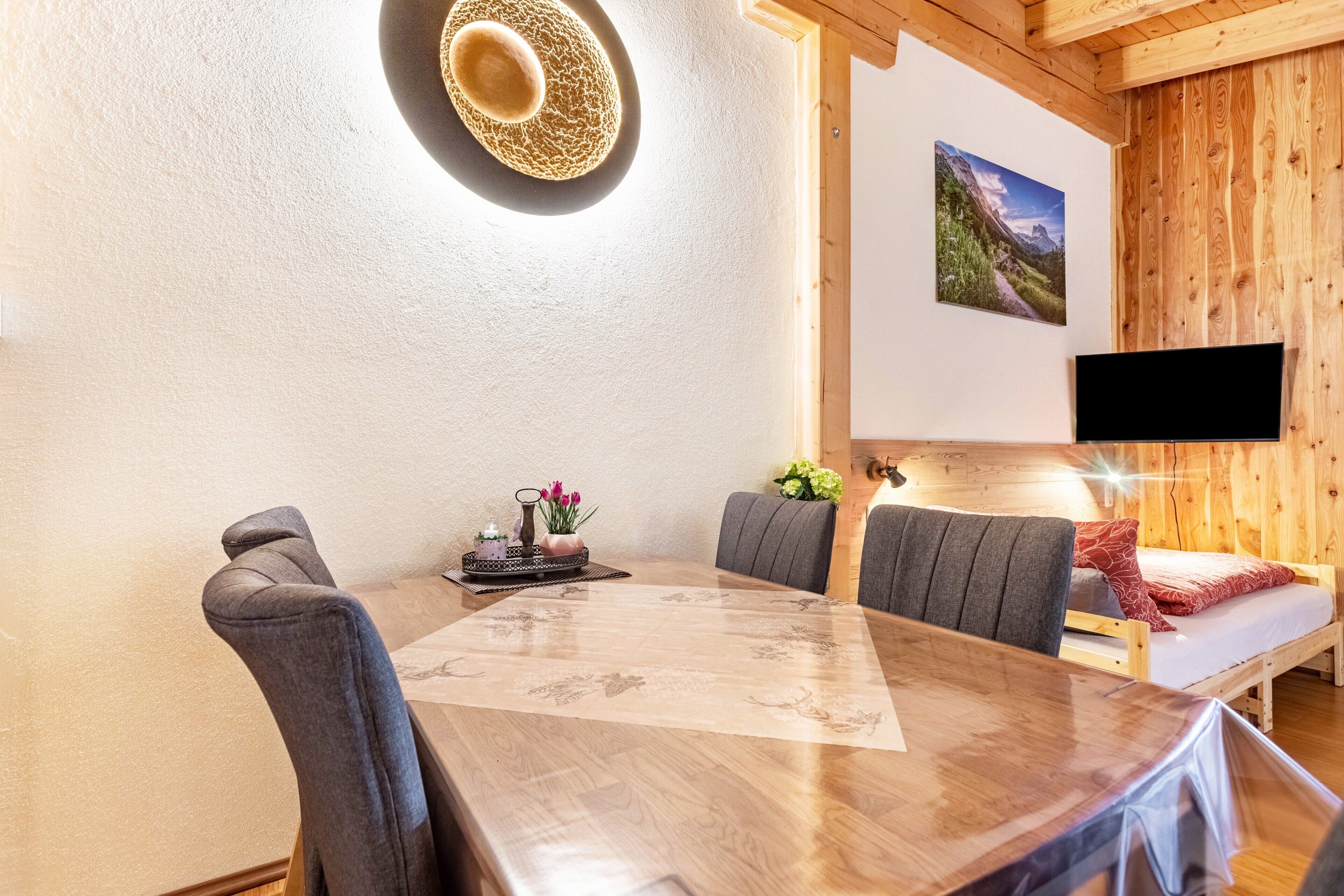Une table à manger en bois avec des chaises, un téléviseur fixé au mur et un lit avec vue sur la montagne.
