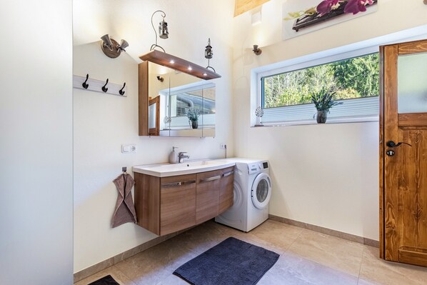 A modern bathroom with a built-in washing machine, a large mirror, and a window with a view of greenery.
