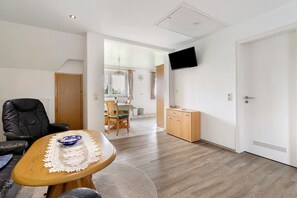 A modern living room with a wooden coffee table, a black leather chair, a dining area with a table and chairs, a kitchen with wooden cabinets, and a mounted TV.