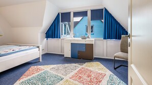 A bedroom with a bed, a desk with a chair, a window with curtains, and a view of a blue house. - Apartment 'Bi Reiner' with Private Terrace and Wi-Fi (Nordstrand)