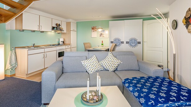 A modern living room with a grey sofa, a coffee table with candles, a kitchen in the background, and a decorative wall with nautical-themed items. - Apartment 'Bi Reiner' with Private Terrace and Wi-Fi (Nordstrand)