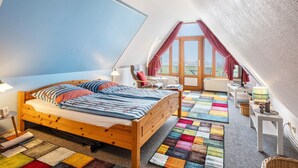 A bedroom with a wooden bed, a colorful patchwork carpet, a small table with a lamp, and a view through a window. - Apartment 'Bi Buje' with Balcony and Wi-Fi (Nordstrand)