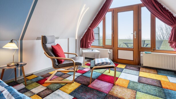 A room with a sloped ceiling, a window with curtains, a chair, a small table, and a colorful carpet. - Apartment 'Bi Buje' with Balcony and Wi-Fi (Nordstrand)