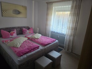 Una cama doble con ropa de cama rosa y blanca, una mesita de noche, un radiador y una ventana con cortinas traslúcidas.