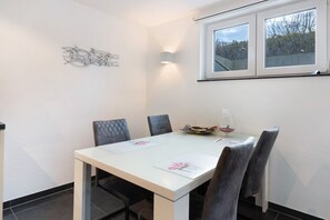 A dining area with a white table, four chairs, a decorative wall piece, and a window with a view of trees. - Goldbecker Apartment with Private Terrace and Wi-Fi (Gaienhofen)