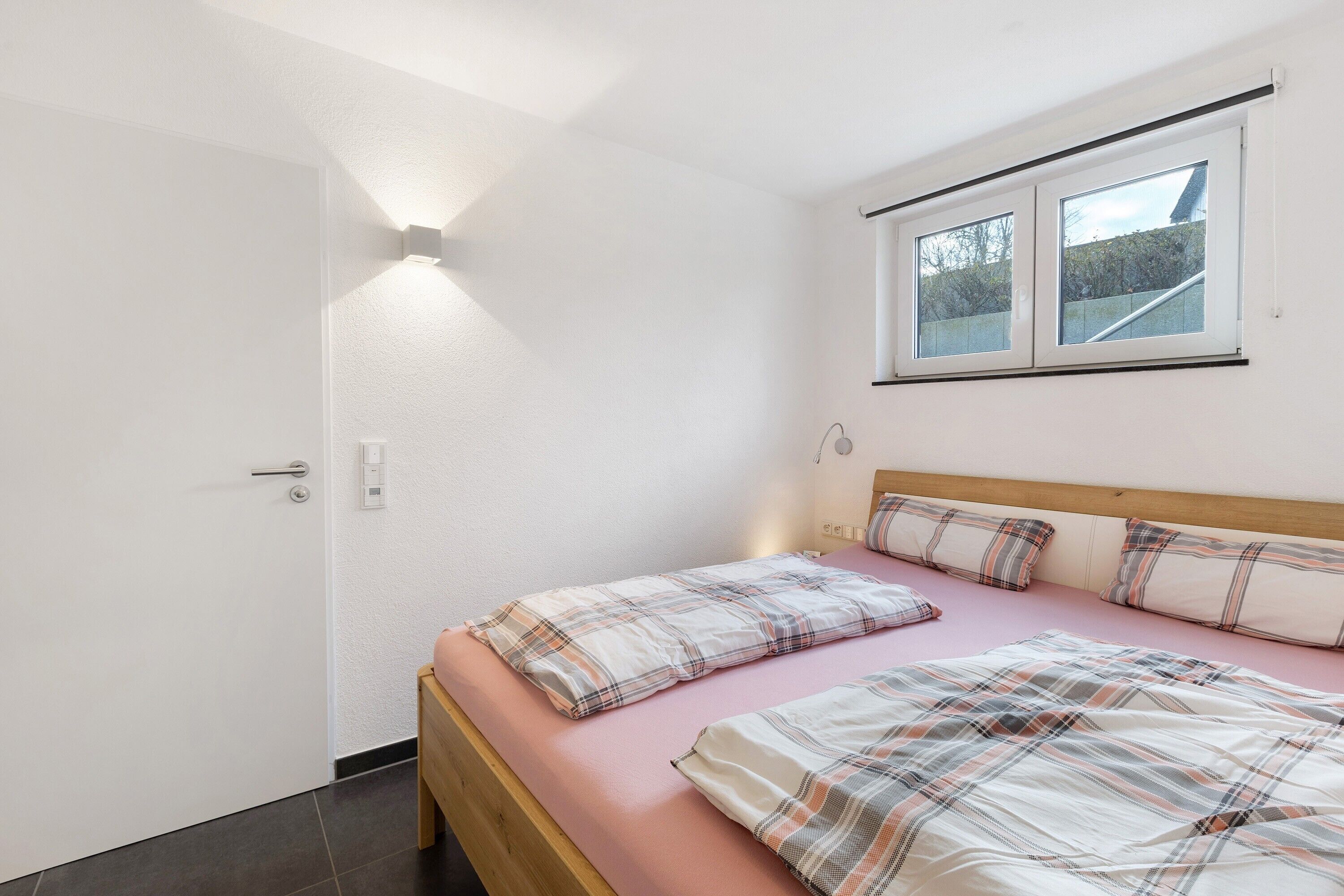 A bedroom with a bed, a window, a door, and a wall-mounted light.