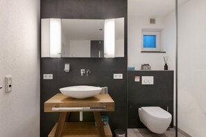 A modern bathroom with a wooden vanity, a white sink, a mirror, a toilet, and a window with a view. - Goldbecker Apartment with Private Terrace and Wi-Fi (Gaienhofen)