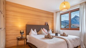 A bedroom with a wooden headboard, a bed with white linens, two bedside lamps, a small table, and a view of mountains through a window. - Design Apartment Au with Mountain View, Balcony and Wi-Fi (Au)