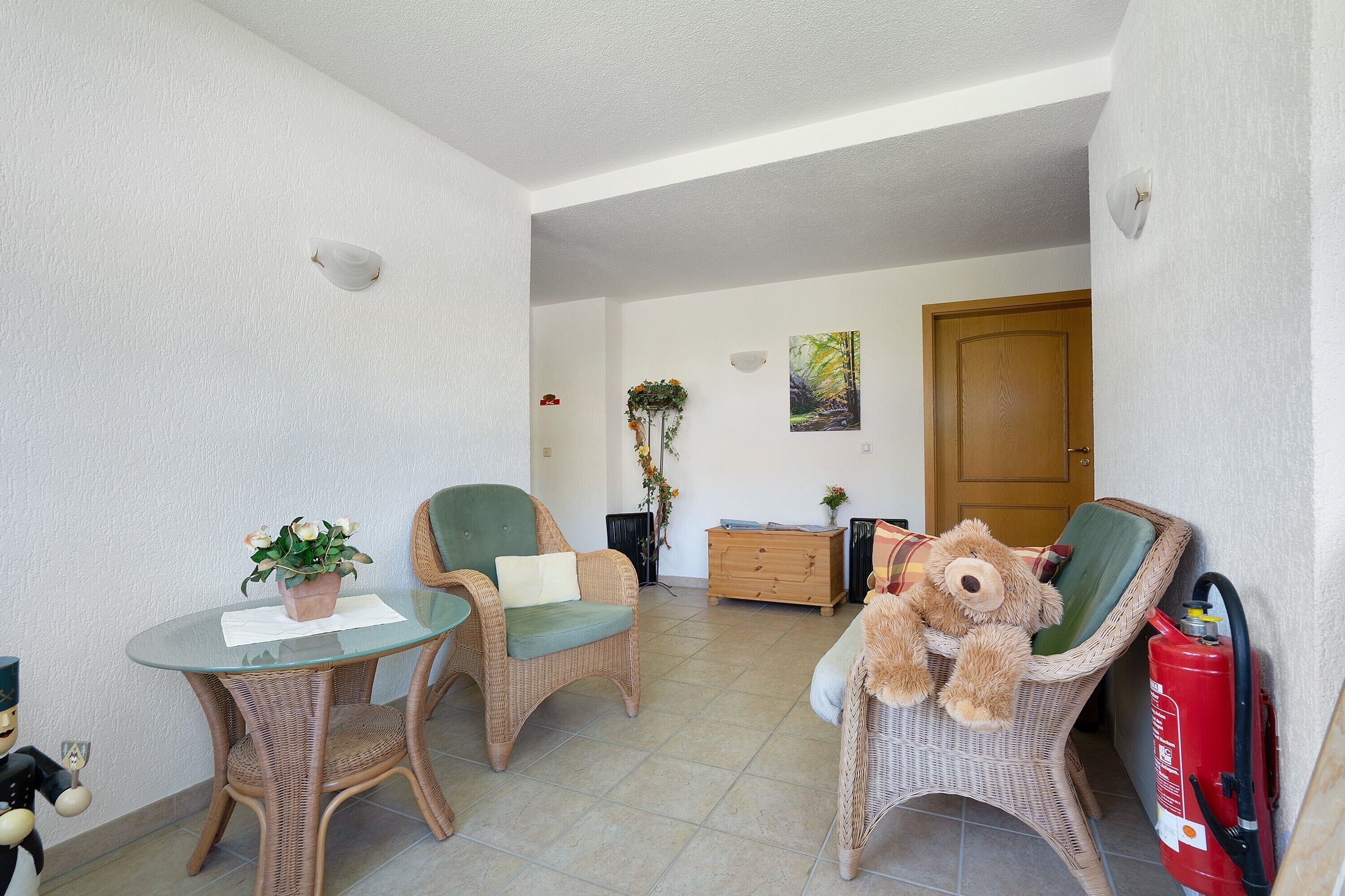 Une pièce meublée de meubles en osier, avec une table en verre ornée d’une plante, un ours en peluche, un extincteur et une porte en bois.