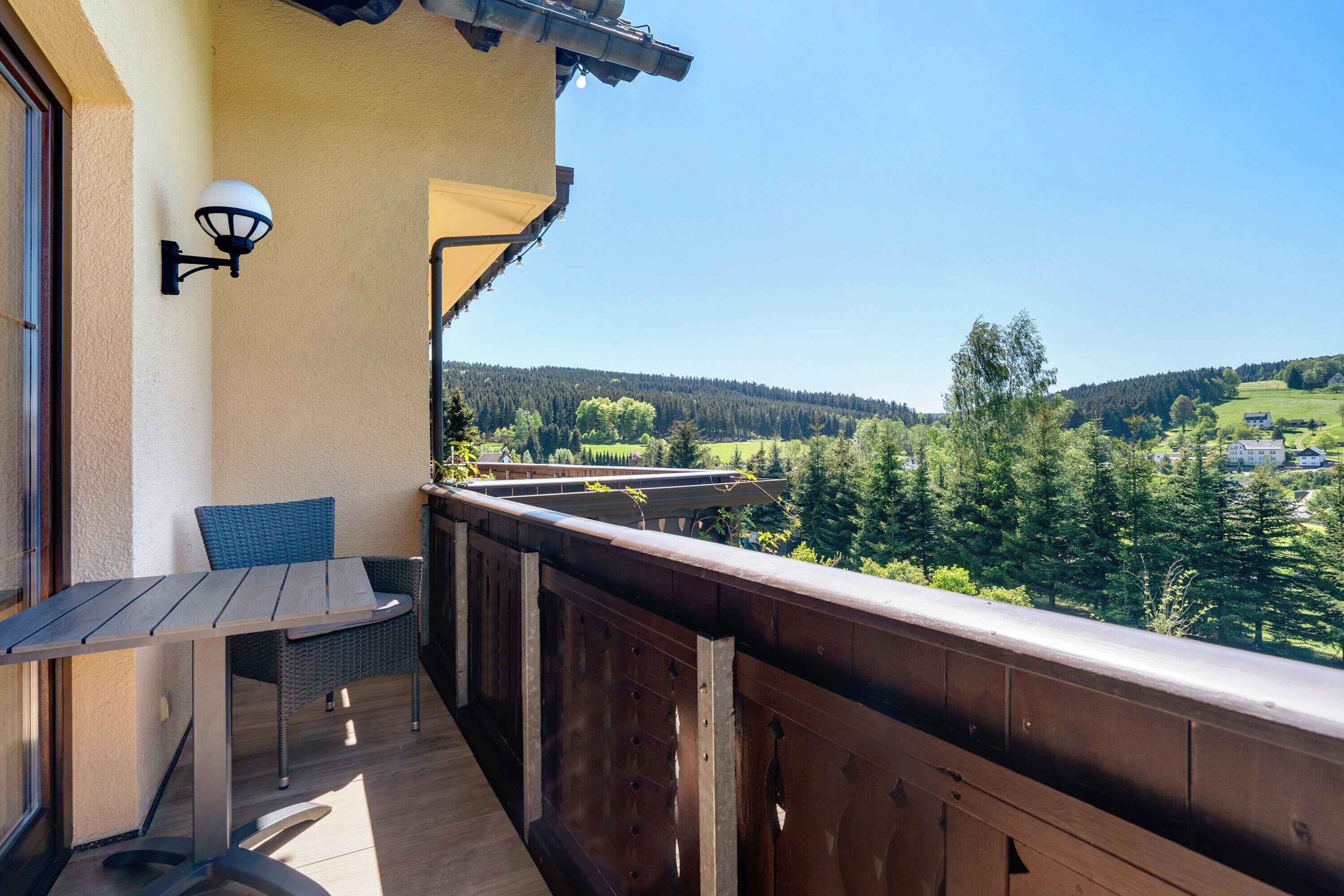 A balcony with a table and chair, a lamp, and a view of a forested landscape.