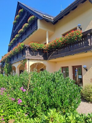 A multi-story building with balconies adorned with red flowers, a welcoming entrance, and a well-maintained garden.
