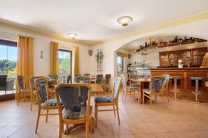 A dining area with wooden chairs and tables, a bar with stools, and a view of greenery outside.
